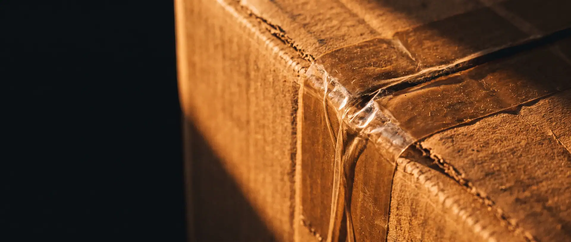 Close view of the corner of a brown cardboard carton sealed with clear packing tape, warm light across the tape and deep shadow on the left