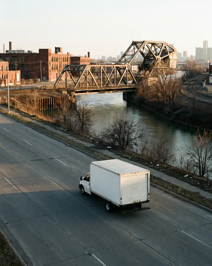 A white box truck on a wide four-lane road beside a river, a steel bascule bridge and brick industrial buildings ahead, rail curves along the bank and a downtown skyline in low gold light