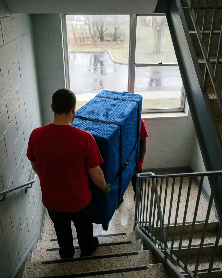 Seen from above and behind, a mover in a red T-shirt carries a tall dresser wrapped in a blue moving blanket down a cinder-block apartment stairwell with steel rails and terrazzo steps, a second mover hidden behind the piece except a red sleeve and a glove, a window onto a wet parking lot ahead
